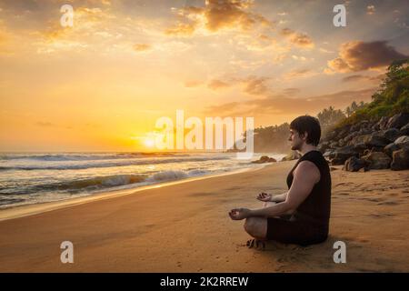 Young sporty fit man doing yoga meditating on tropical beach Stock Photo