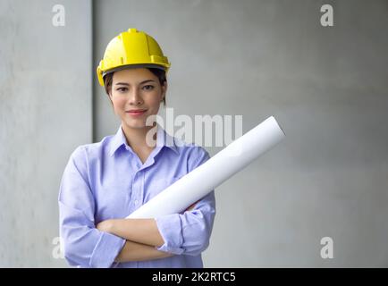 Young woman in construction helmet and floor plan stand with arms crossed in front of gray plaster wall. Work environment of engineers at the construction site of housing projects. Stock Photo