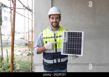 Young engineer in hardhat safety vest and glove pointing finger at solar cell panel, present a source of energy to generate direct current electricity with a smile. Stock Photo