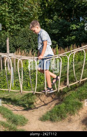 Cheerful blond boy walks on rope bridge on a wooden playground in a public park. Stock Photo