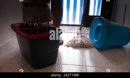 mop wring into the bucket, ready to clean the floor with tiles and colorful curtain on the door in the background.second mop overturned to the side. Stock Photo