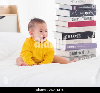 baby little girl toddler looking at a stack of books on bedroom white bed Stock Photo