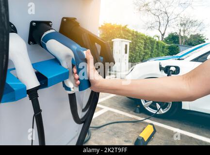 Hand holding electric plug-in charger on blur car charging. EV car charging at electric car charging station. EV car charging point. Electric vehicle fast charge. Sustainable power. Green energy. Stock Photo