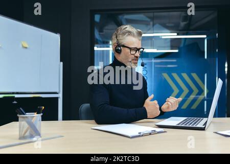 Senior experienced gray-haired businessman with headset, using laptop for online meeting, video call with colleagues businessman mature working inside modern office building. Stock Photo