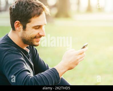 Young happy man sitting in the park and using smartphone. Stock Photo