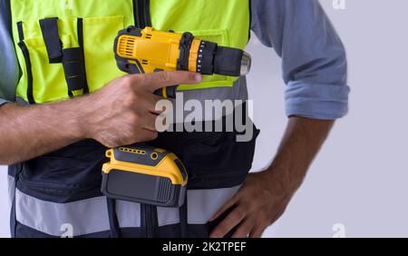 A yellow cordless electric drill with battery in the hand of a construction worker wearing an green reflective safety vest. Close up Stock Photo