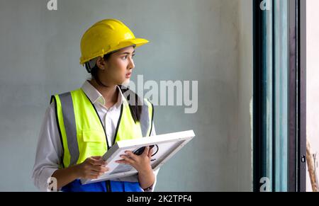 Young asian woman in a safety vest and hardhat holding solar cell panel while looking out of the window. Work environment of engineers at the construction site. Stock Photo