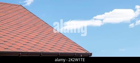 New roof with red shingles against the blue sky. Tiles on the roof of the house. Use to advertise roof fabrication and maintenance. Spotted texture. Affordable roofing. Banner with copy space. Stock Photo