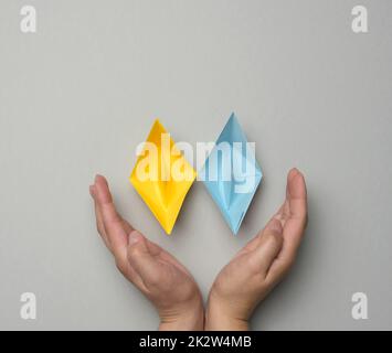 Two yellow-blue paper boats in female palms on a gray background Stock Photo