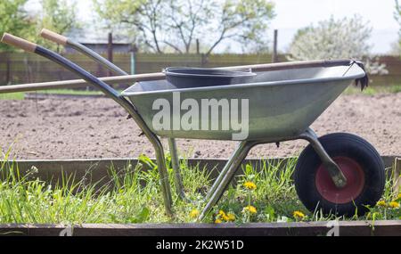 Gray metal garden wheelbarrow with two handles and one wheel. The wheelbarrow is in the garden or garden. Gardener's wheelbarrow in the backyard. Garden cleaning. Stock Photo