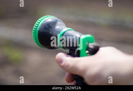 The gardener's hand holds a hose with a sprayer and waters the plants in the garden. Green atomizer in hand with a large lan. Horticulture hobby concept. Watering with a hose. Stock Photo