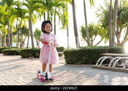 Happy Asian little kid girl wear safe helmet playing pink kick board on road in park outdoors on summer day Stock Photo