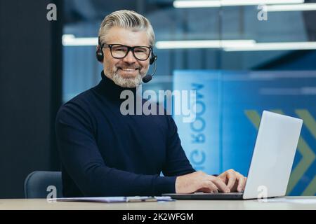 Portrait of mature gray haired businessman, man in glasses smiling and looking at camera, senior boss with headset for video call using laptop, executive working inside modern office building. Stock Photo