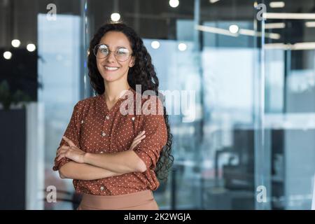Young beautiful and successful hispanic businesswoman smiling and looking at camera, female worker working inside office wearing glasses and curly hair, arms crossed portrait Stock Photo