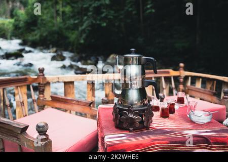 front view closeup of traditional Turkish tea chrome kettle and glasses served on a restaurant table with chairs near a river in an outdoor setting Stock Photo