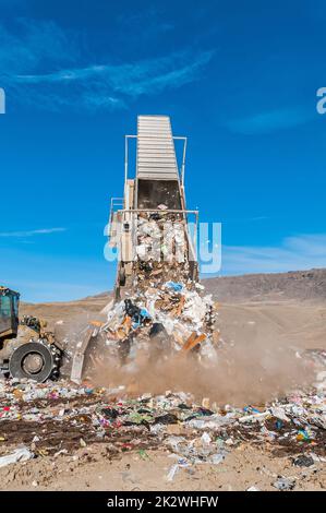 A trailer from a semitrailer truck is in a landfill tipper being raised and dumps out trash, while a soil compactor bulldozer pushes the trash. Stock Photo