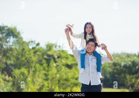 father and carrying an excited girl on shoulders having fun and enjoying outdoor lifestyle together playing aircraft toy Stock Photo