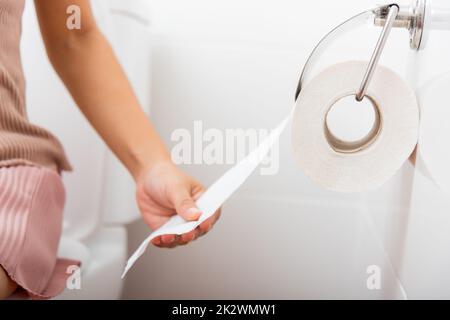 Closeup hand pulling toilet paper roll in holder for wipe.Closeup hand pulling toilet paper roll in holder for wipe, woman sitting on toilet she taking and tearing white tissue on wall to towel clean in bathroom, Healthcare concept Stock Photo