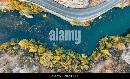 A road on a mountain slope overlooking a river with colorful vegetation and trees on the edge of the slope. Drone flight view. Background Stock Photo