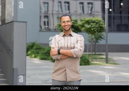Young successful businessman smiling and looking at camera with crossed arms, man outside office building wearing shirt and glasses, startup investor. Stock Photo