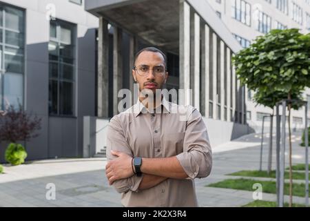 Serious and pensive young office worker with crossed arms looking at camera, businessman in shirt outside office building pensive. Stock Photo