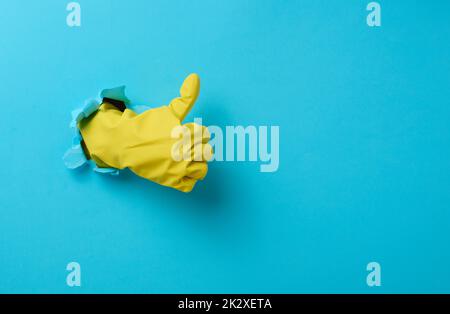 hand in a yellow household rubber glove sticks out of the torn hole and shows a gesture okay, thumb up. Blue background Stock Photo
