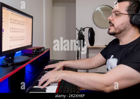 young latin caucasian man with a beard, glasses and headphones, sitting playing the piano and singing, studying digital sheet music with the computer Stock Photo