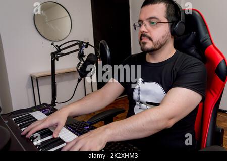 front side view of a young latin man, with a beard, glasses and headphones sitting at home concentrated studying music online and playing the piano Stock Photo