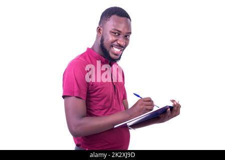 portrait of handsome happy young man wearing red t-shirt, writing in notebook with pen while smiling at camera. Stock Photo