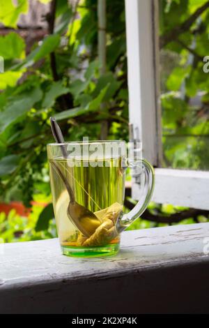 Tea and tea bag under the summer sun. A tea bag in a glass transparent mug on the windowsill and against the background of green summer trees. Stock Photo