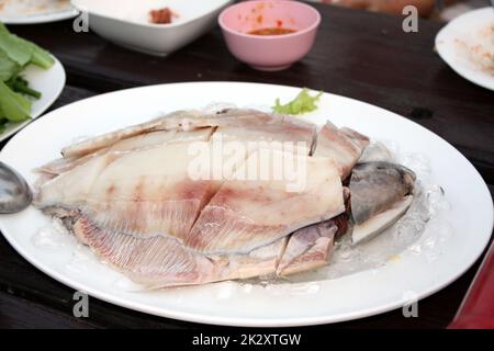 Fresh Gray pomfret, Chinese silver pomfret fish cut into pieces arranged in a white plate topped with ice. On the wooden table in the restaurant, prepare for the shabu pot. Selective focus. Stock Photo