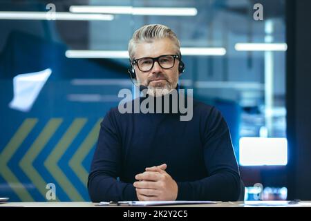 Video call, serious and mature businessman looking thoughtfully at camera, gray-haired man using headset for remote communication and online meeting. Stock Photo