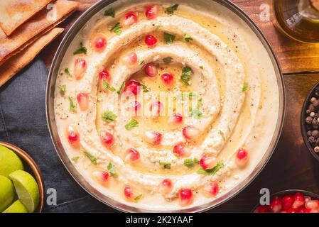 Arabic cuisine; Traditional creamy hummus dip in dark bowl. Topped with olive oil, fresh pomegranate seeds and parsley. Top view with close up. Stock Photo