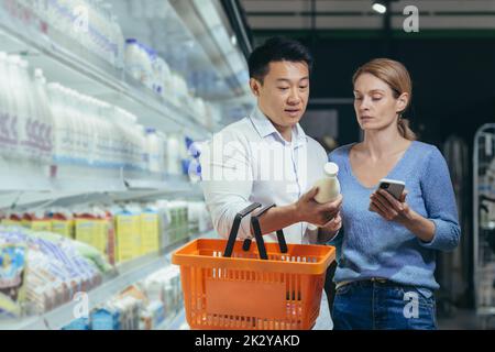 Young happy Asian couple using smartphone in supermarket with shopping cart choosing products while grocery store. browsing smart phone joyful. daily food shopping list people make a choice pick Stock Photo