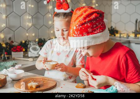Little dark-haired girl 3 years old and boy 8 years old in red Santa hat decorate gingerbread cookies with icing. Siblings in white Christmas Stock Photo