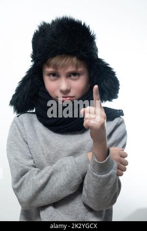 Winter hat with earflaps and scarf, portrait of a boy in winter clothes, the boy holds up his index finger. Stock Photo