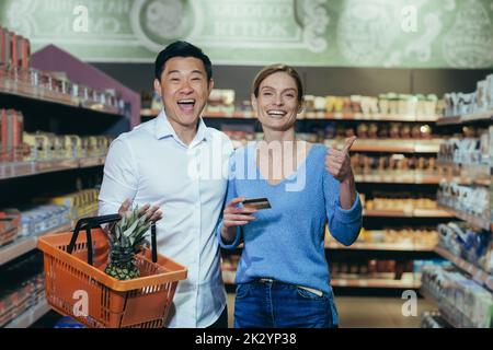 Happy diverse family couple man and woman shoppers in supermarket, looking at camera and smiling happy shopping, holding phone and bank credit card in hand, with shopping basket. Stock Photo