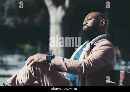 A portrait of an elegant confident bald black man with a well-groomed beard enjoying the sun while sitting on a park bench; a side view of a bearded A Stock Photo