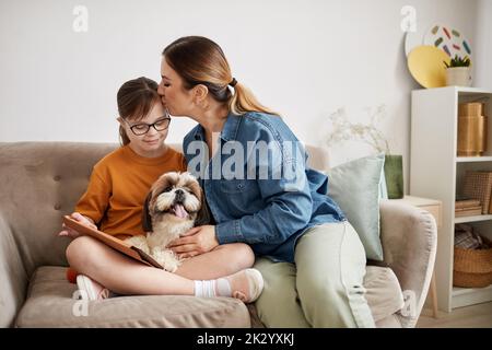 Minimal portrait of loving mother kissing daughter with Down syndrome while sitting on couch at home Stock Photo