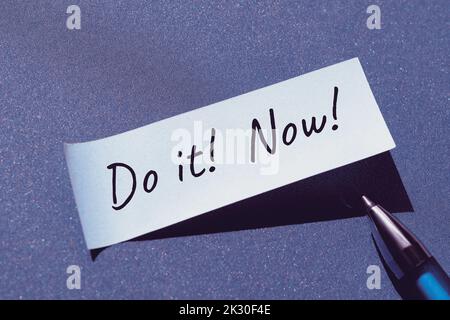 Do it now motivator message written on sticky paper, office sticker with pen. Single simple minimal motivation note stiker on blue paper background. Stock Photo