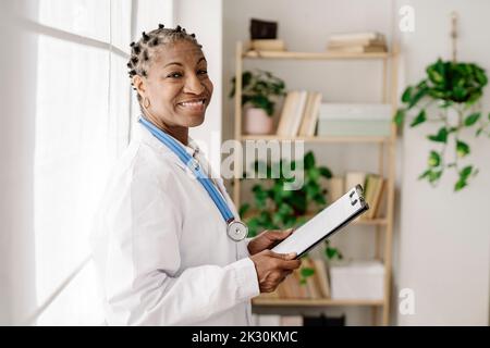 Smiling female doctor with clipboard standing by window at home office Stock Photo