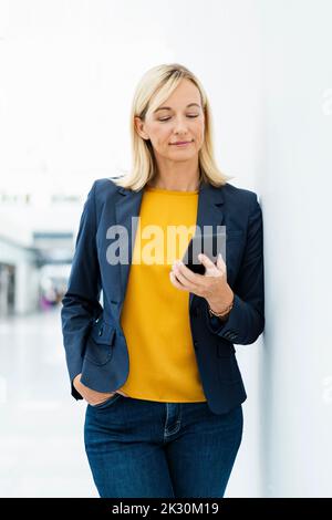 Smiling businesswoman using phone standing by wall Stock Photo