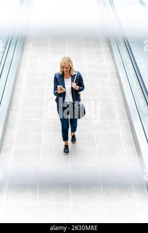 Smiling businesswoman using phone walking on walkway Stock Photo