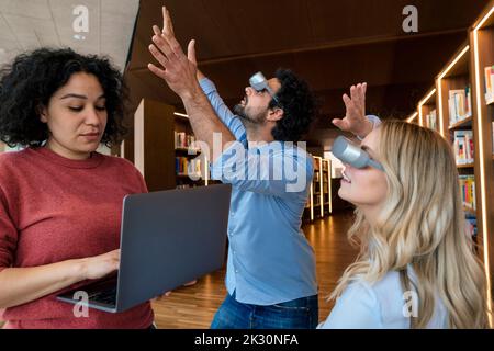 Woman working on laptop by colleagues with VR glasses gesturing in library Stock Photo