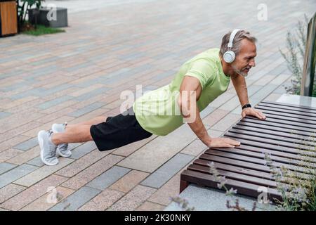 Man wearing wireless headphones doing push-ups on bench Stock Photo