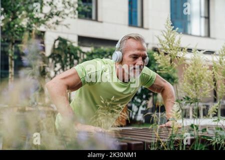 Mature man wearing wireless headphones doing push-ups on bench Stock Photo
