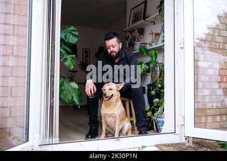 Depressed man holding drink stroking dog sitting on stool at doorway Stock Photo