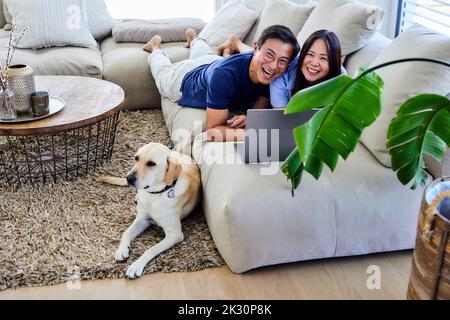 Cheerful couple with laptop lying on sofa in living room at home Stock Photo