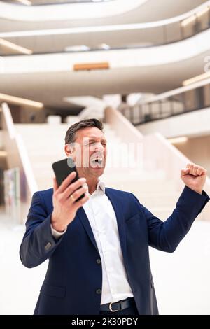 Cheerful businessman with smart phone clenching fist in lobby Stock Photo
