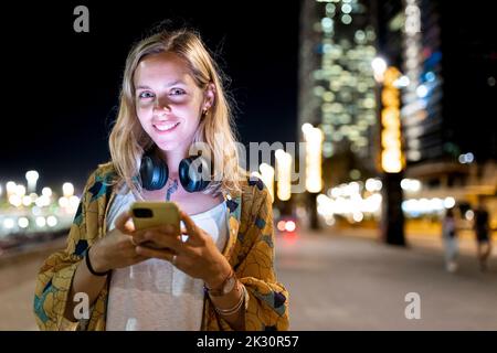 Happy young blond woman wearing wireless headphones holding mobile phone at night Stock Photo